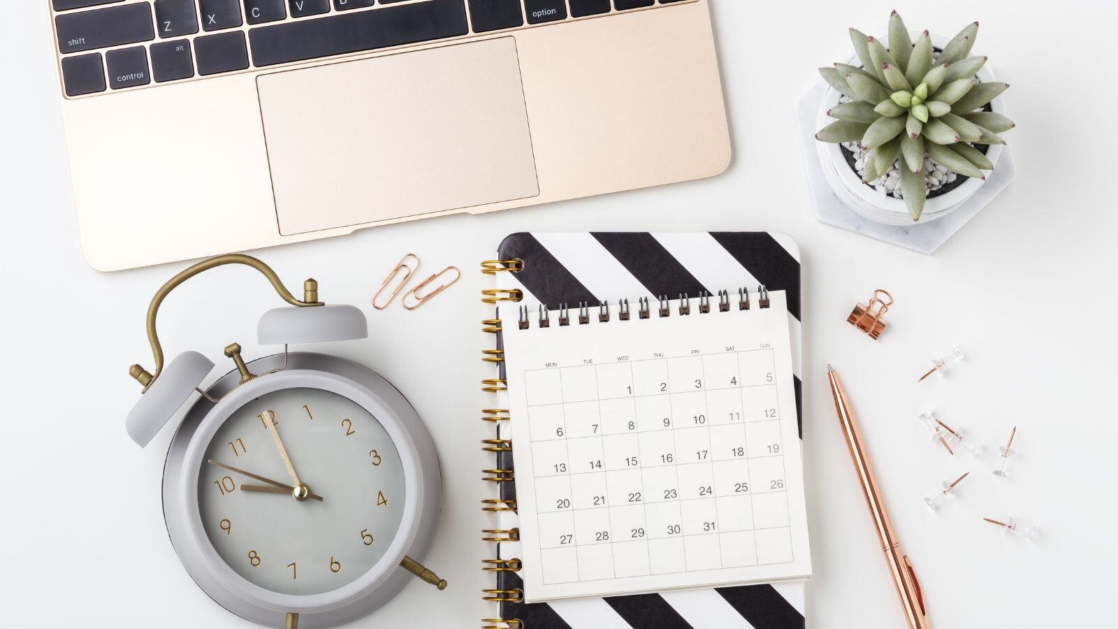 A laptop keyboard positioned above a clock, a paper calendar, and some pens