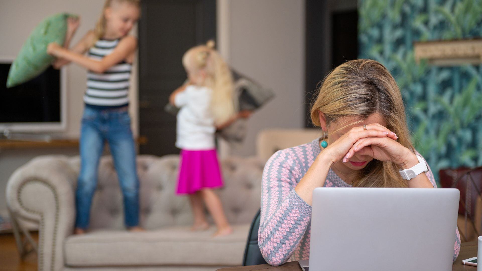 Mom is trying to work on her laptop while her two daughters are bouncing on the couch behind her