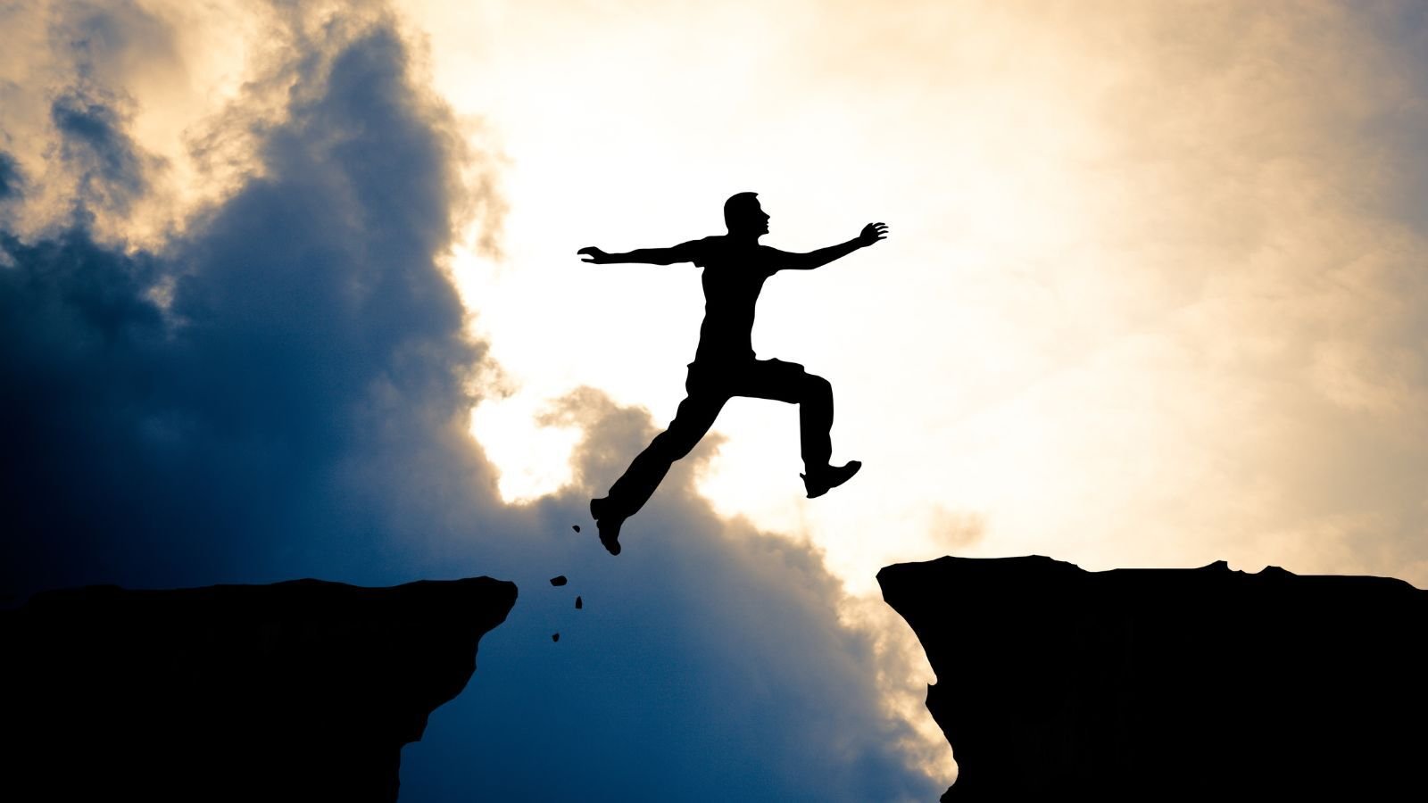A man jumping from one rock cliff to another in front of an open sky with big clouds